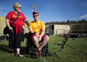 Marine veteran Cpl. Richard Stalder smiles with sunglasses on and looking at the camera while seated in his wheelchair outside on a sunny day with his archery kit. He is accompanied by his mother, who is standing to his left, with sunglasses on and looking at him.