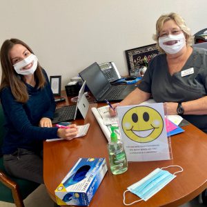 Two women wearing clear masks sit at a desk.