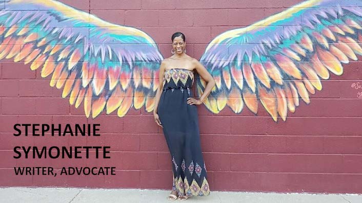 Photo of black woman standing in front of a wall with multi-colored wings painted on it.