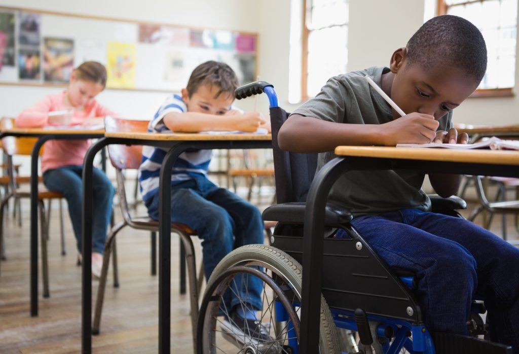 Three students write at their desks. The boy in the front is sitting in a wheelchair.
