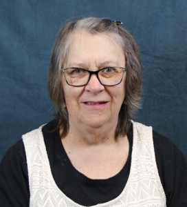 A white woman with short grey hair and glasses. She wears a white vest over a black shirt.